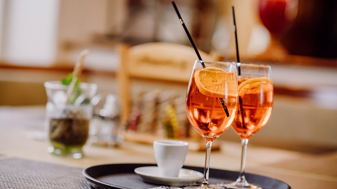 Zwei Gläser mit orangefarbenem Getränk stehen auf einem Schaumstofftablett. Im Hintergrund sind ein kleiner Kaffeebecher und eine Glasvase mit Kräutern zu sehen. | © Gasthof Landseele