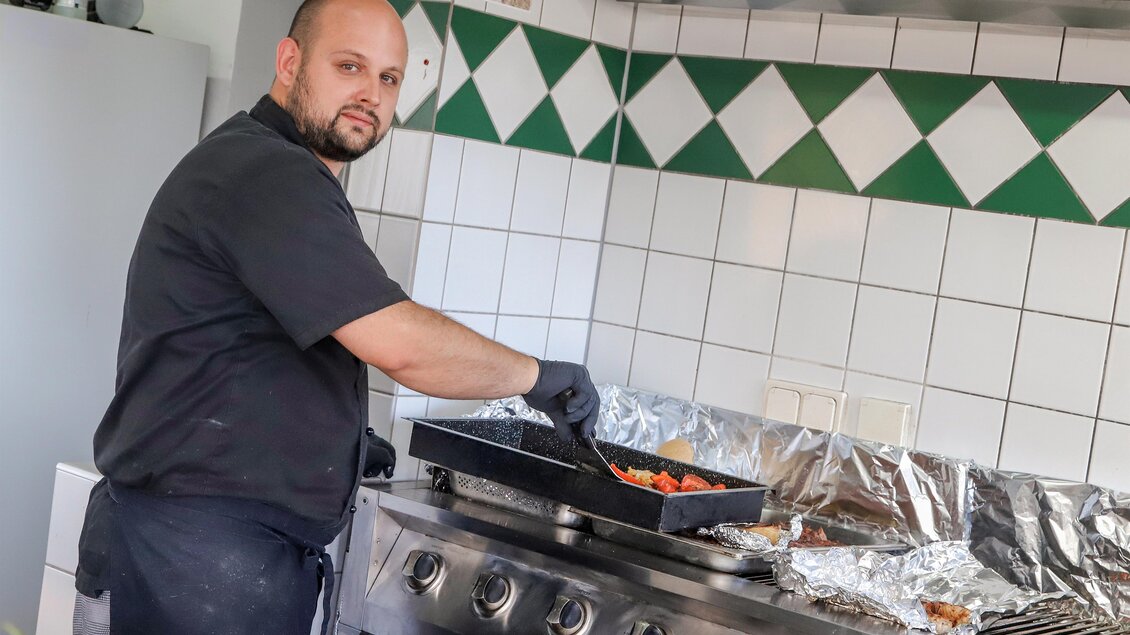 Ein Koch bereitet frische Zutaten in einer modernen Küche vor. Er schaut konzentriert in die Kamera, während er mit einer Pfanne arbeitet. | © Iris Bloder