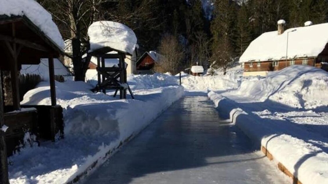 Ein gefrorener Kanal, umgeben von Schnee und Bäumen. Im Hintergrund sind traditionelle Holzhäuser zu sehen. | © Gasthof Leitner