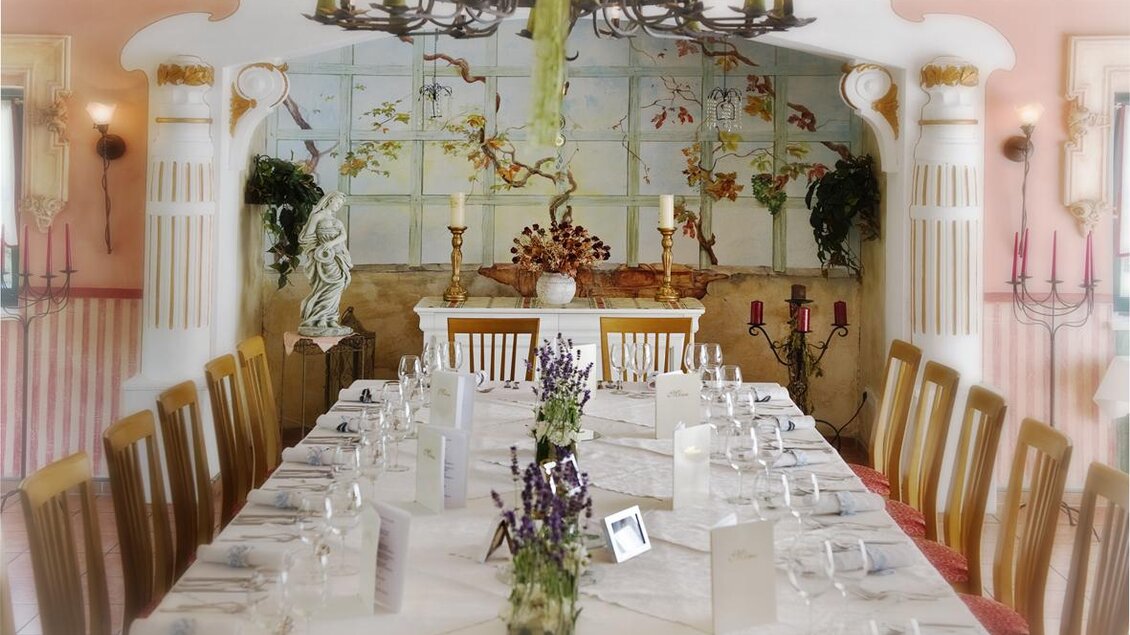 Ein eleganter Speisesaal mit einem langen Tisch und dekorativen Blumenarrangements. Die Wände sind mit kunstvollen Malereien und Säulen verziert. | © Gasthof Lobmingerhof