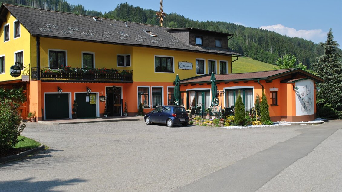 Ein buntes Gasthaus umgeben von grünen Hügeln. Der Parkplatz ist leer und die Sonne scheint. | © Gasthof Lobmingerhof