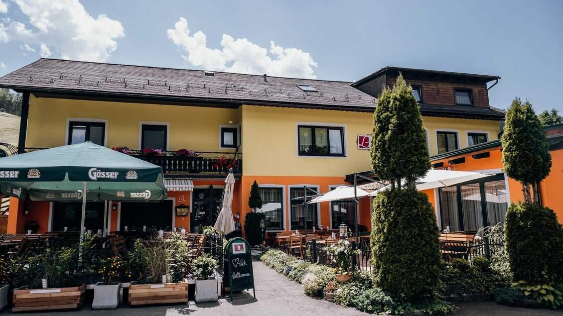 Ein charmantes Restaurant mit einer gelben Fassade und gemütlichen Sitzgelegenheiten im Freien. Die Umgebung ist von Pflanzen und Bäumen umgeben, ideal für eine entspannte Atmosphäre. | © Gasthof Lobmingerhof