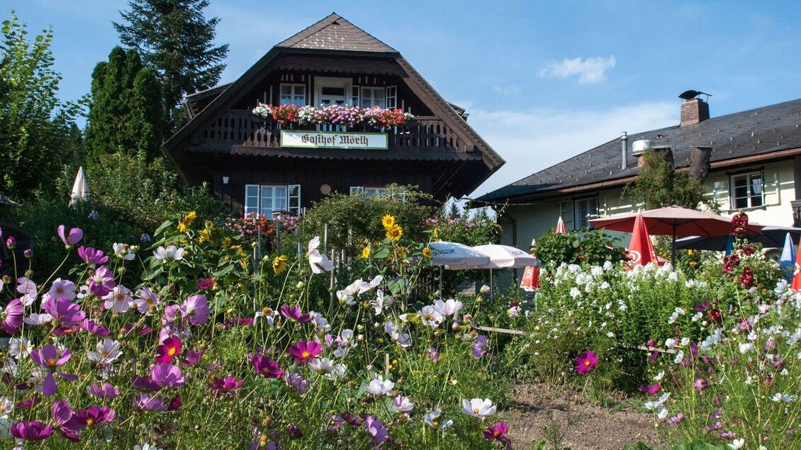 Ein charmantes Haus mit einem Balkon, umgeben von bunten Blumen und einem schönen Garten. Im Hintergrund sind weitere Gebäude und ein blauer Himmel zu sehen. | © Gasthof Mörth