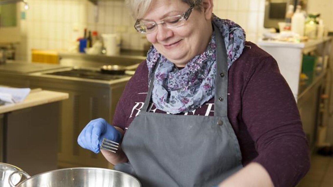 Eine Frau in einer Küche bereitet Zutaten in großen Schüsseln vor. Sie trägt eine Schürze und Handschuhe und lächelt dabei. | © Stephan Friesinger
