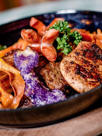 An artfully arranged plate with grilled meat, colorful vegetables, and crispy chips. The dish is presented in a black bowl on a wooden board. | © TV Region Graz - Mias Photoart