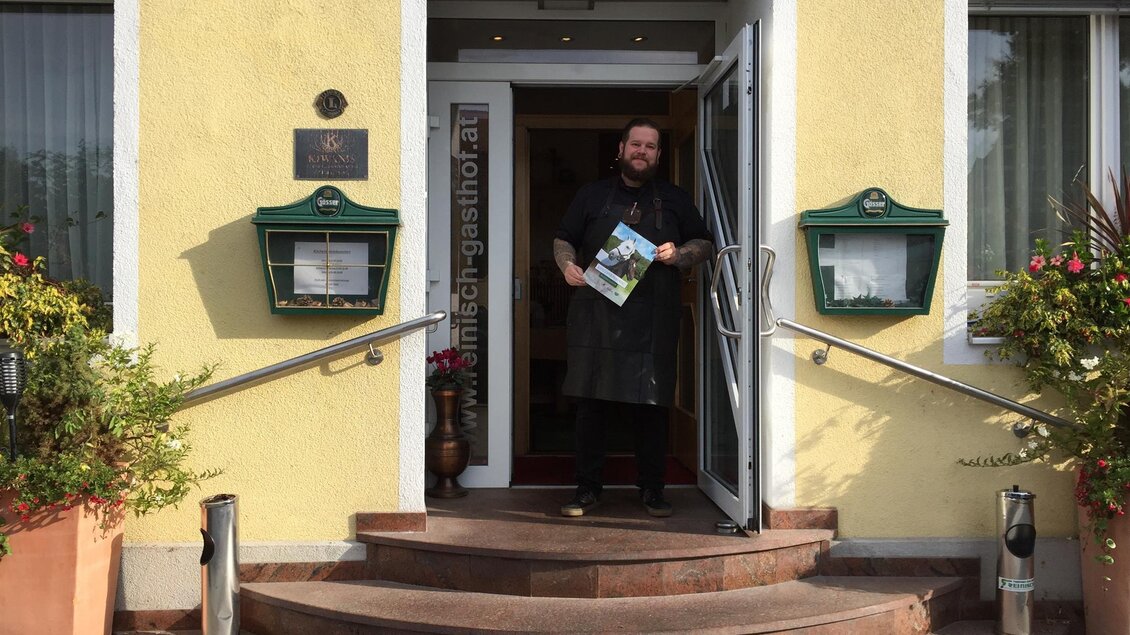 Gastgeber steht im Eingang des Gasthofs Reinisch, hält Prospekt vor gelber Fassade mit Blumentöpfen in Köflach.  | © TV Lipizzanerheimat