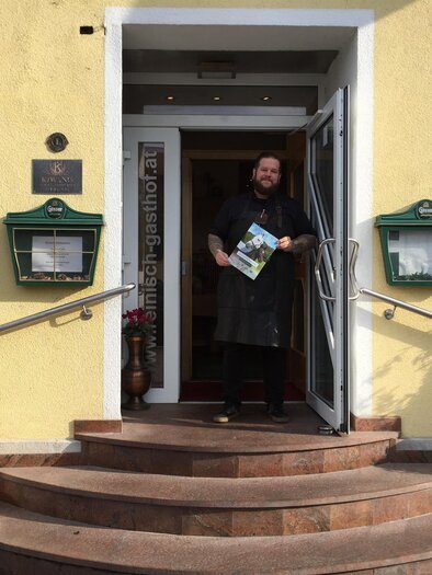 Gastgeber steht im Eingang des Gasthofs Reinisch, hält Prospekt vor gelber Fassade mit Blumentöpfen in Köflach.  | © TV Lipizzanerheimat