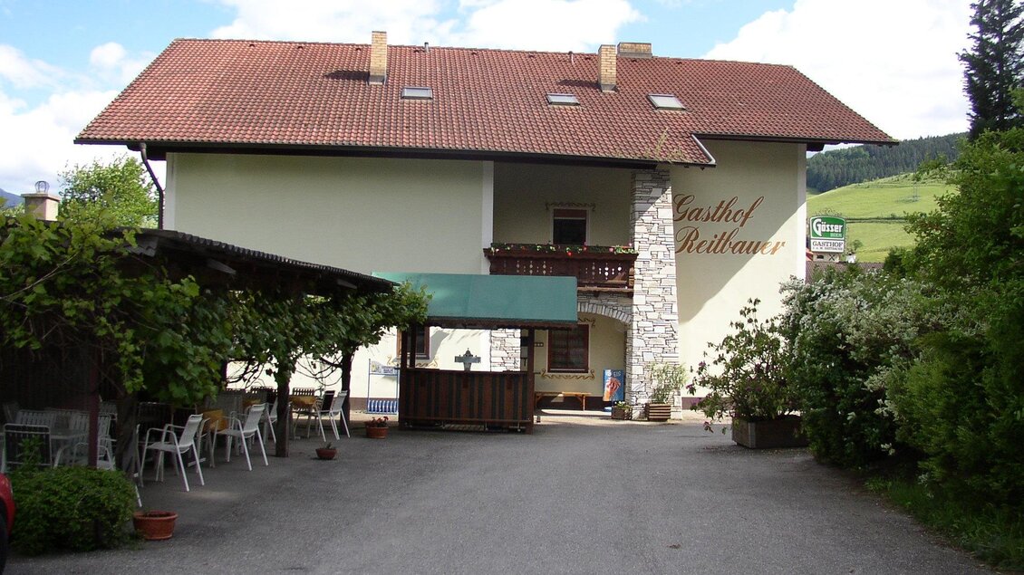 Ein gemütliches Gasthaus mit einem großen, überdachten Außenbereich. Umgeben von Bäumen, bietet es eine einladende Atmosphäre in ländlicher Umgebung.