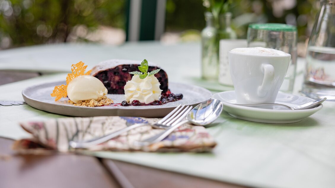 Ein köstliches Stück Kuchen mit Beeren, Sahne und einer Kugel Eis. Daneben steht eine Tasse Kaffee auf einem Tisch. | © 5komma5sinne