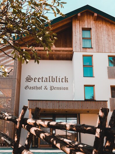A modern guesthouse with wood paneling and an inviting entrance. Surrounded by trees and a fence, it appears friendly and welcoming. | © Gasthof und Pension Seetalbllick