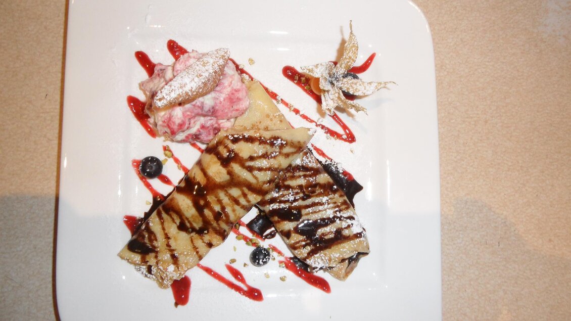 Ein appetitlicher Teller mit zwei gefüllten Crepes, dekoriert mit Schokoladensauce und Beeren. Dazu gibt es eine Portion Erdbeereis und eine essbare Blüte. | © SAM