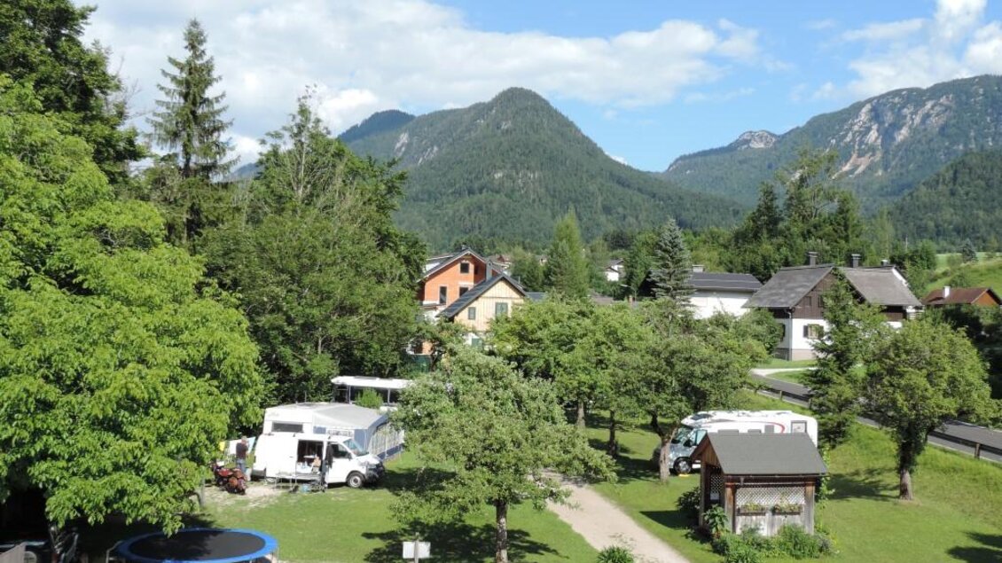Eine idyllische Landschaft mit Bergen im Hintergrund und einem Campingplatz. Es gibt Wohnmobile, einen Parkplatz und einen Trampolin inmitten von Bäumen. | © Gasthof Staud'nwirt
