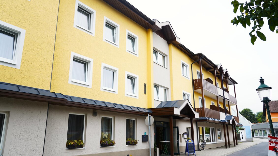 Ein modernes mehrstöckiges Gebäude in gelber Farbe mit Fenstern und Balkonen. Die Straße vor dem Gebäude ist gepflastert und hat einige Grünflächen. | © TV Hochsteiermark/Amelie Kertelits