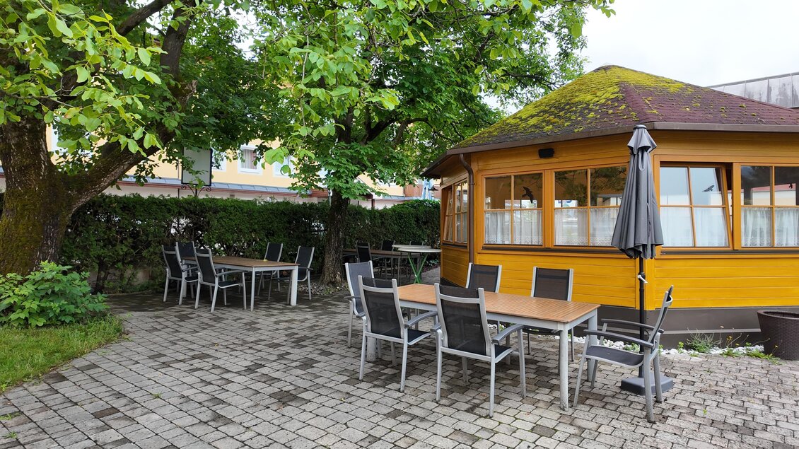 Ein gemütlicher Außenbereich mit Holztischen und Stühlen. Im Hintergrund steht ein gelbes Häuschen unter einem Baum. | © TV Hochsteiermark/Amelie Kertelits