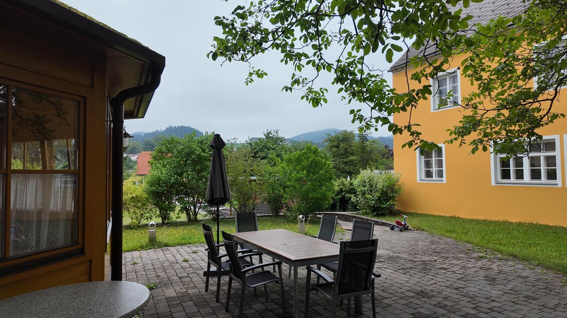 Ein ruhiger Garten mit einem großen Tisch und mehreren Stühlen. Im Hintergrund ist ein gelbes Haus und grüne Bäume zu sehen. | © TV Hochsteiermark/Amelie Kertelits