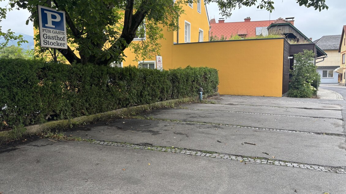 Ein Parkplatz mit einer grünen Hecke und einem Schild für das Gasthaus Stöckl. Im Hintergrund sind einige Gebäude zu sehen. | © TV Hochsteiermark/Amelie Kertelits