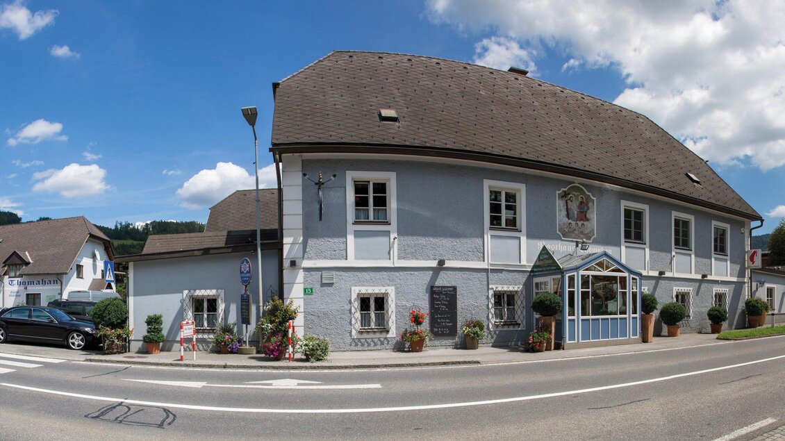 Ein traditionelles Gebäude an einer Straße mit blauer Fassade und einem Einfahrtstor. Es gibt Blumen und ein paar Autos in der Nähe. | © TV Region Graz - Lunghammer