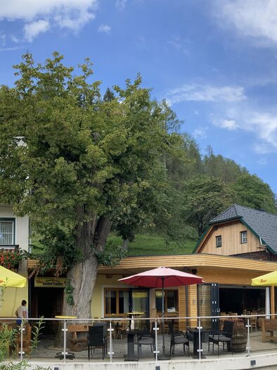 Ein charmantes Gasthaus mit mehreren Etagen und Balkonen. Im Freien stehen bunte Sonnenschirme für Gäste. | © Gasthof Trattner