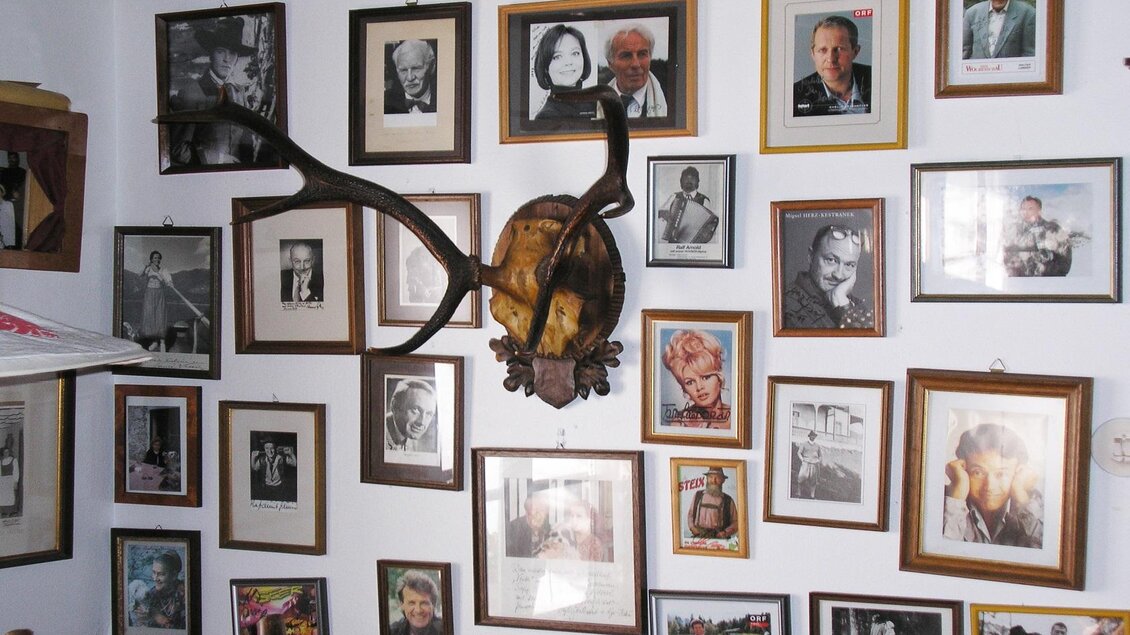 Eine Wand voller gerahmter Fotos und Portraits, die verschiedene Personen zeigen. Darüber hängt ein Geweih, das einen rustikalen Akzent verleiht. | © Gasthaus Veit