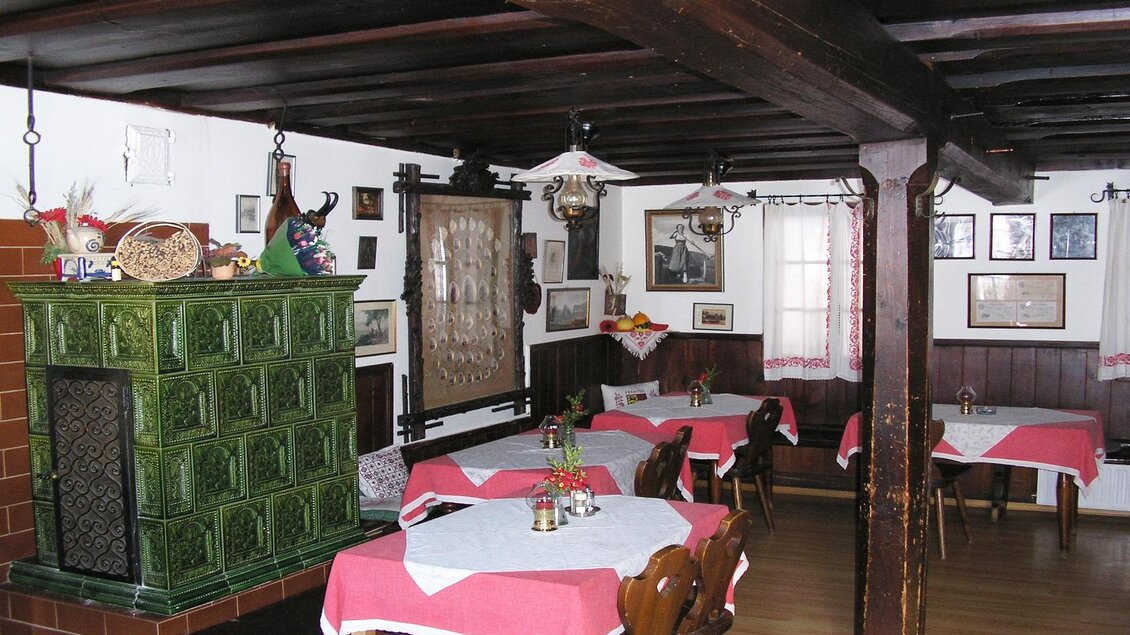 Ein gemütliches Restaurant mit rustikalem Holzinterieur und dekorativen Bildern an den Wänden. Es gibt Tischdecken in Rot und Weiß sowie einen klassischen Kachelofen. | © Steinegger