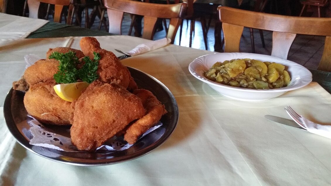 Ein Teller mit knusprig frittiertem Hähnchen, garniert mit einer Zitronenscheibe und Petersilie. Nebenan liegt eine Schüssel mit Beilagen aus Gurken und Kartoffeln. | © Gasthaus Walch