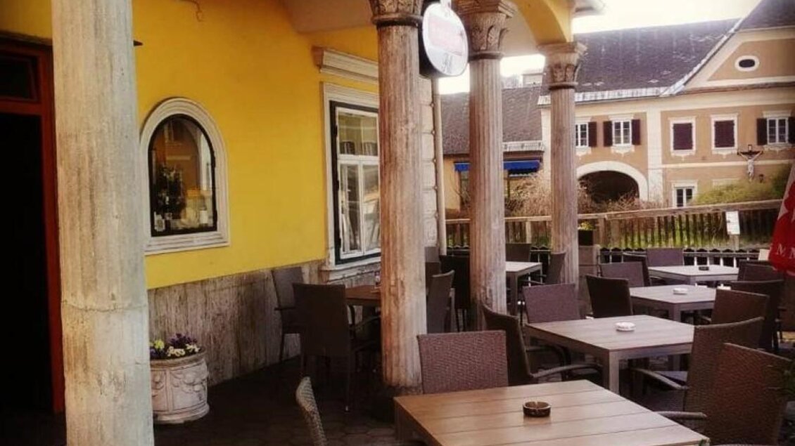 Ein einladender Außenbereich eines Cafés mit Holztischen und Stühlen. Die Wände sind gelb gestrichen und von Säulen umrahmt. | © Gasthof zur Goldenen Krone