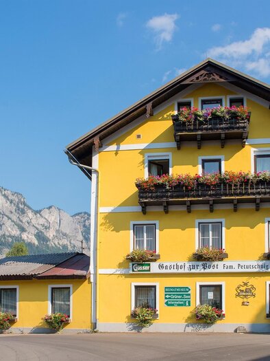 Ein gelbes Gebäude mit Balkonen, geschmückt mit Blumen. Im Hintergrund sind die Berge und ein klarer Himmel zu sehen. | © Gasthof zur Post
