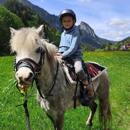 Ein Kind sitzt auf einem grauen Pony in einer grünen Wiese. Im Hintergrund sind Berge und ein blauer Himmel zu sehen. | © Melanie Gonzales