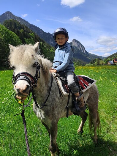 Ein Kind sitzt auf einem grauen Pony in einer grünen Wiese. Im Hintergrund sind Berge und ein blauer Himmel zu sehen. | © Melanie Gonzales