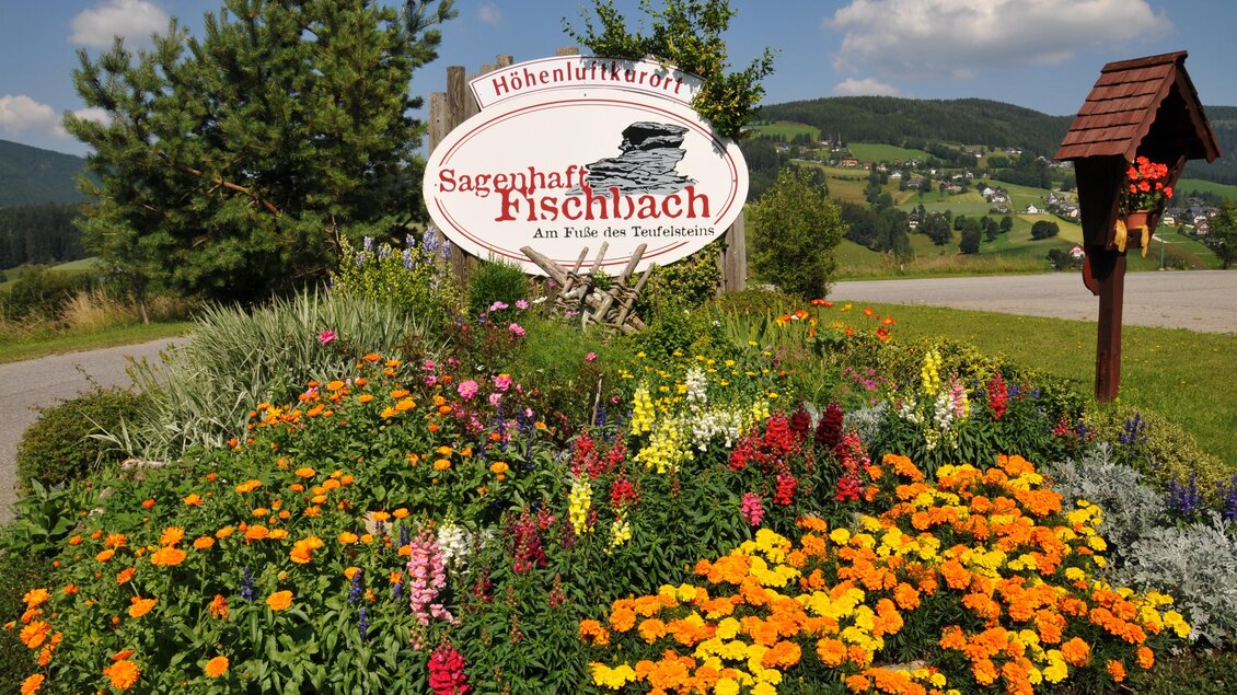 zu sehen ist die Ortstafel mit Marterl und Blumen im Hintergrund der Ort Fischbach. | © Tourismus Fischbach