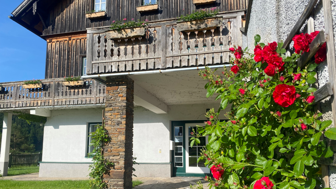 Ein hübsches Holzhaus mit einem Balkon und Blumenarrangements. Die Fassade ist freundlich und einladend, umgeben von grünem Rasen. | © Bettina Scheck