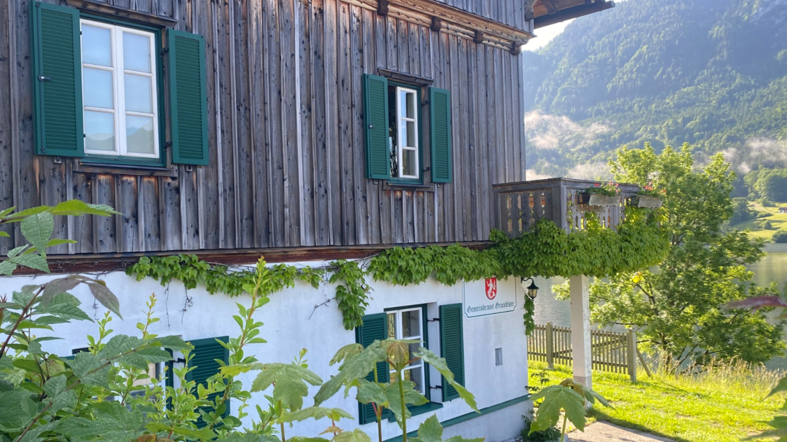 Ein traditionelles Holzhaus mit grünen Fensterläden und einem Balkon. Im Hintergrund sind Berge und eine grüne Landschaft sichtbar. | © Bettina Scheck