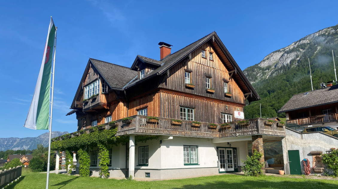 Ein traditionelles Holzhaus in einer malerischen Umgebung. Die Fassade ist mit bunten Blumenkästen geschmückt, umgeben von grünen Wiesen und Bergen. | © Bettina Scheck