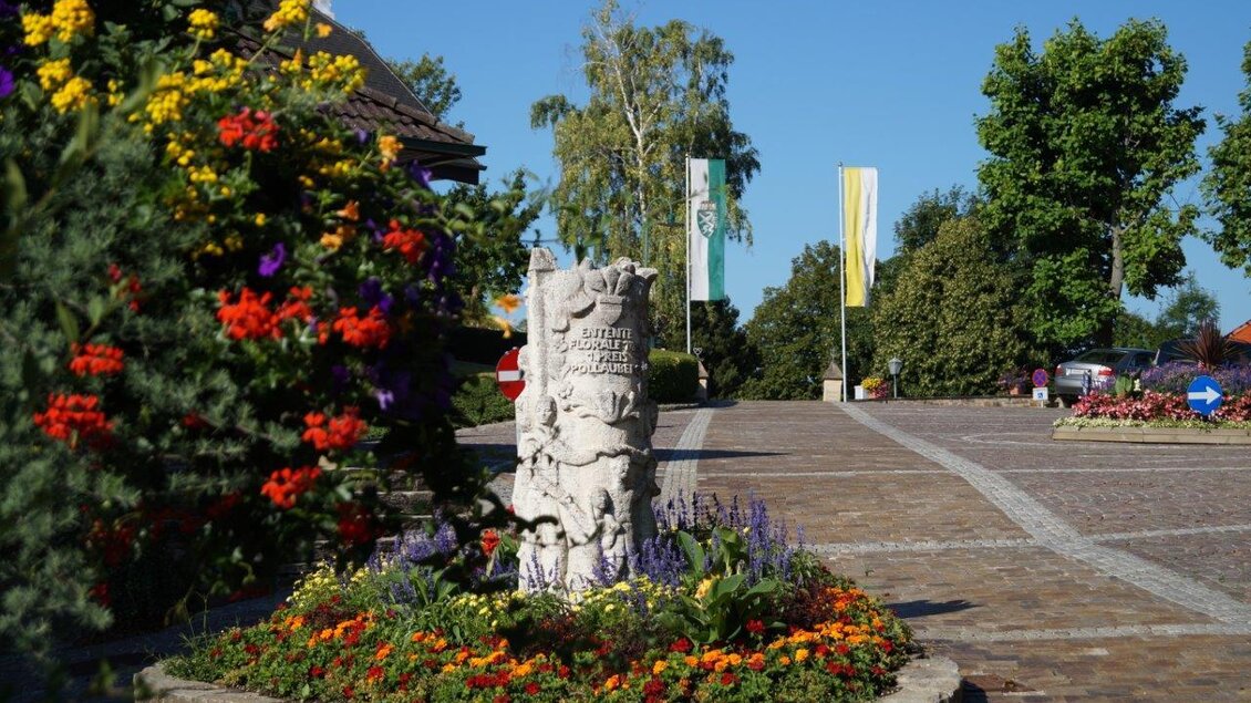 Ein hübscher Platz mit bunten Blumen und einer Steinpfeiler-Skulptur. Im Hintergrund sind Bäume und einige Fahnen zu sehen. | © Gemeinde Pöllauberg