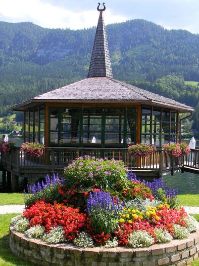 Ein schöner Pavillon am Wasser, umgeben von bunten Blumenbeeten. Im Hintergrund sind grüne Berge und ein klarer, blauer Himmel zu sehen. | © Gemeinde Grundlsee