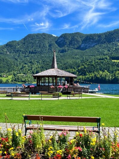 <p>Der schöne Gemeindepark am Grundlsee mit dem Muisikpavillon und blühenden Blumen. Im Hintergrund sind grüne Berge und ein blauer Himmel zu sehen.</p> | © Tourismusverband Ausseerland