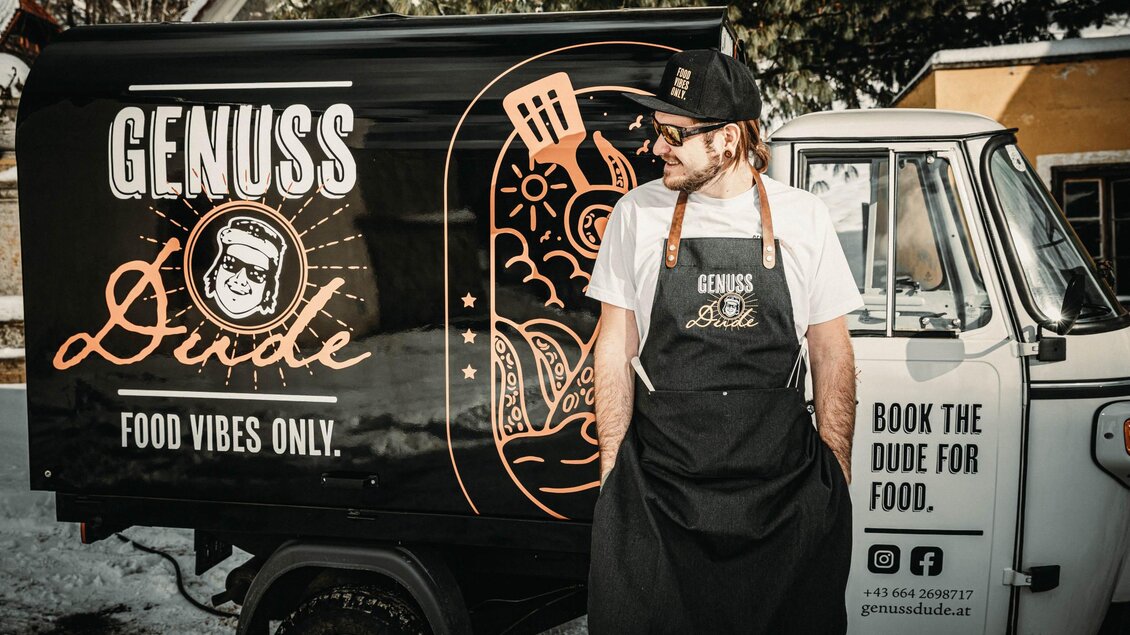 Ein foodtruck mit der Aufschrift „Genuss Dude“. Der Verkäufer trägt eine Schürze und bietet lokale Speisen an. | © Helmut Niemand