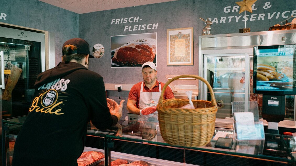 Ein Fleischerei-Laden mit ausgehängtem Fleisch und einem Kunden, der mit dem Verkäufer spricht. Im Hintergrund sind frische Fleischprodukte und ein Korb sichtbar. | ©  www.maxmenkphoto.com