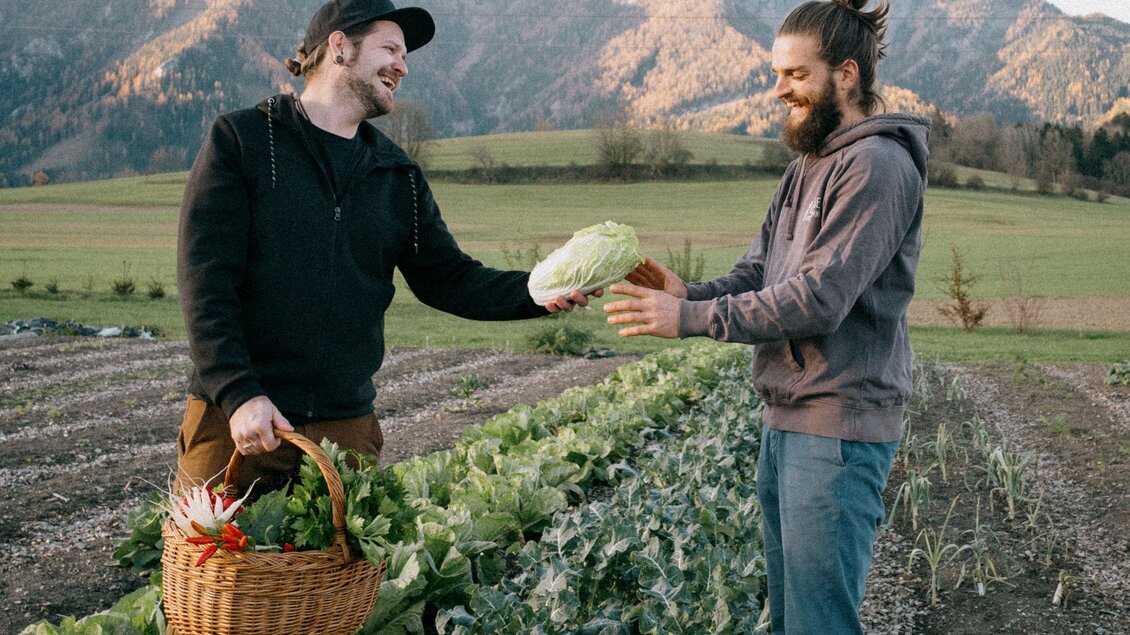 Zwei Männer stehen in einem Gemüsefeld und tauschen frisches Gemüse aus. Im Hintergrund sind Berge und eine grüne Landschaft zu sehen. | ©  www.maxmenkphoto.com