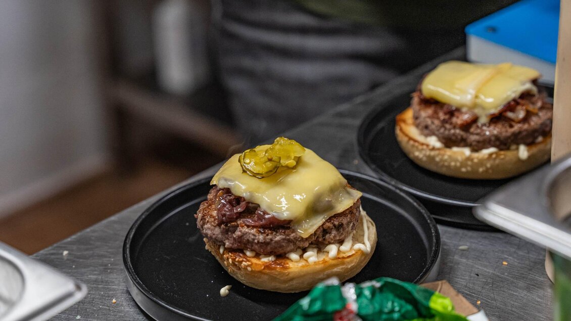 Ein saftiger Burger auf einem Brötchen mit geschmolzenem Käse und Gurken. Die Burger liegen auf einem schwarzen Teller in einer Küche. | © Christoph Hofer
