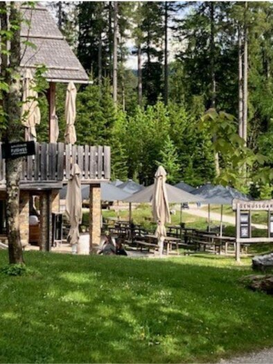 Der Genussgarten ist ein gemütlicher Imbissplatz umgeben von Bäumen. Inmitten einer grünen Wiese am See  stehen Tische unter Sonnenschirmen. | © TVB Ausseerland Salzkammergut (c) Heike Temmel