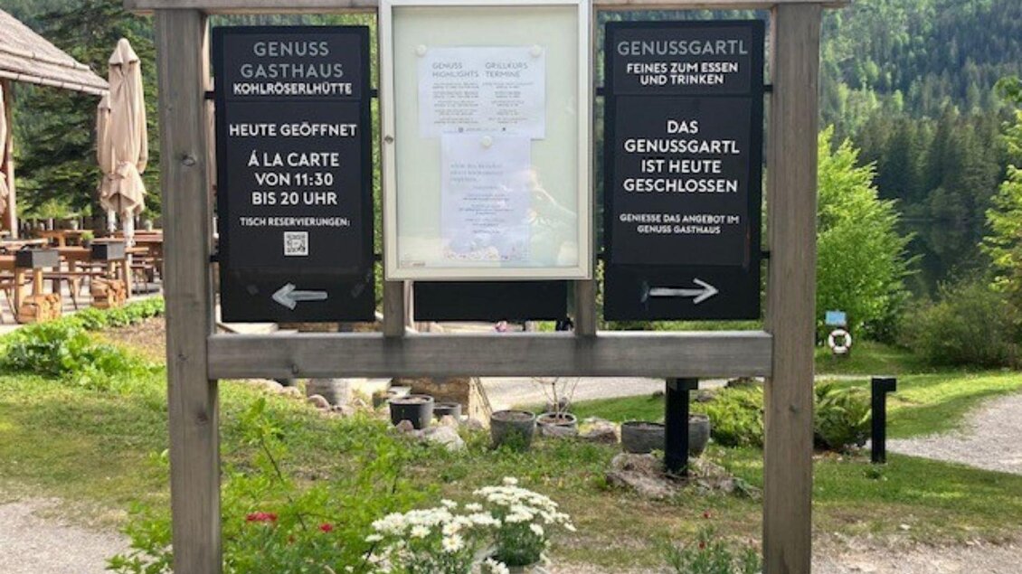 <p>Der Wegweiser ist ein Holzschild mit Informationen über das Restaurant und die Öffnungszeiten linker Hand und das Genuss Gartl mit Öffnungszeiten rechter Hand. Im Hintergrund sind Bäume und eine grüne Landschaft sichtbar. | © TVB Ausseerland Salzkammergut (c) Heike Temmel