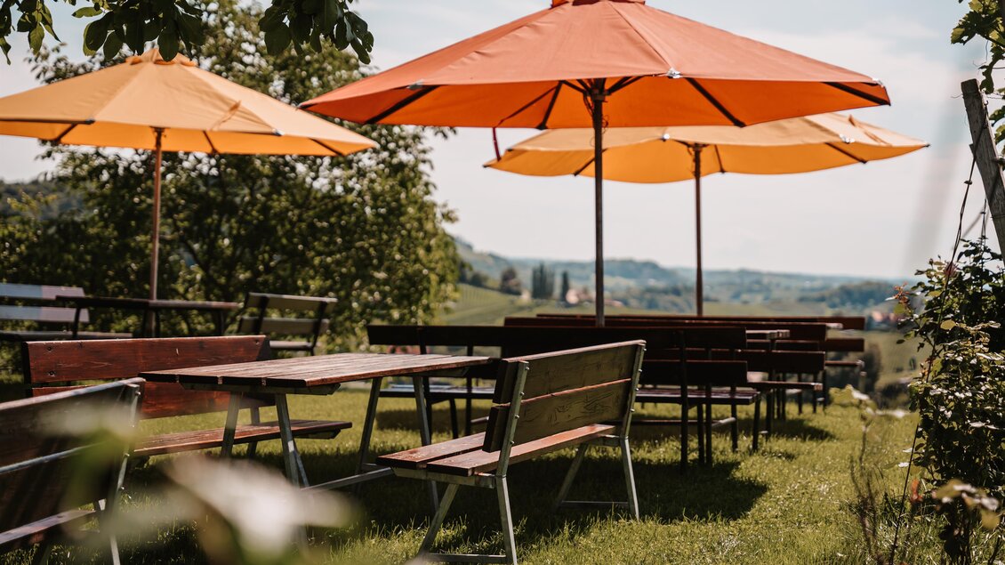 Ein schöner Garten mit Holztischen und orangenen Sonnenschirmen. Im Hintergrund ist eine sanfte Hügellandschaft zu sehen. | © Genuss-Geschwister Kästenburg