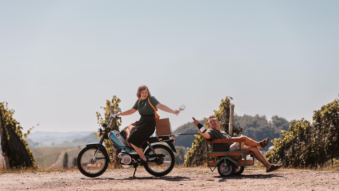 Eine Frau fährt auf einem Moped und winkt fröhlich. Neben ihr sitzt ein Mann entspannt in einem Schubkarren, umgeben von Weinreben. | © Genuss-Geschwister Kästenburg