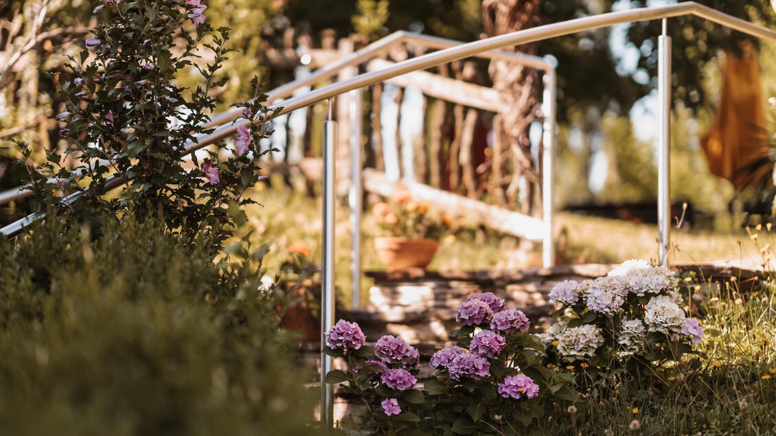 Eine schöne Gartenansicht mit blühenden Pflanzen und einem Handlauf, der zu einer Treppe führt. Die natürliche Umgebung ist idyllisch und einladend. | © Genuss-Geschwister Kästenburg