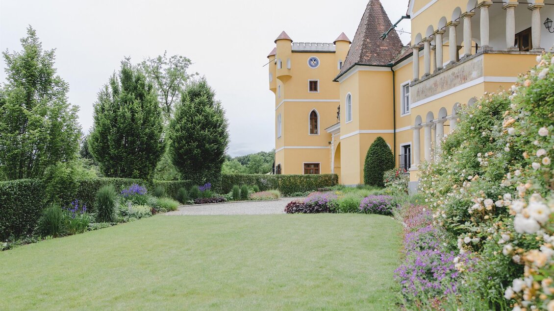 Ein wunderschönes Anwesen mit einem gelben Gebäude und einem Turm. Der Garten ist grün und blühend, umgeben von Bäumen und Blumen. | © Georgi Schloss | Oliver Haring