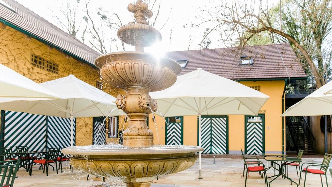 Ein wunderschöner Brunnen in einem Innenhof mit Sonnenschirmen. Im Hintergrund sind bunte Türen und Bäume zu sehen. | © Oliver Haring