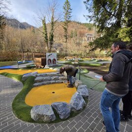 An outdoor mini-golf course with colorful lanes and natural stones. Two people are watching a player as he positions his ball.