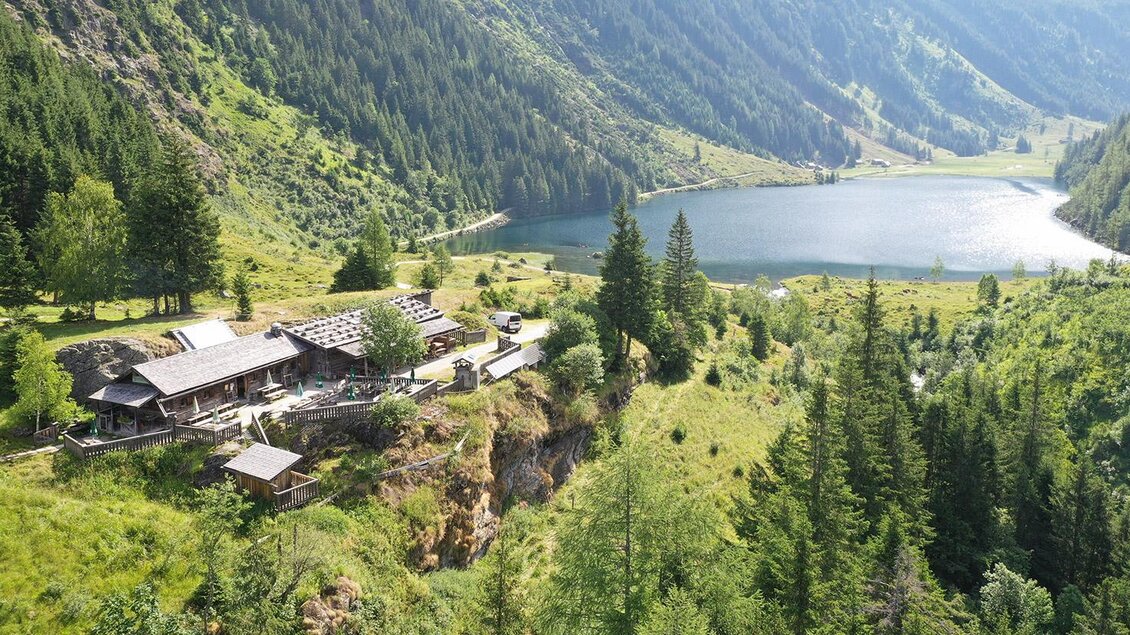 Eine malerische Landschaft mit einem See und einem Bauernhaus umgeben von Bäumen. Die Berge im Hintergrund vervollständigen das idyllische Bild. | © Herbert Raffalt