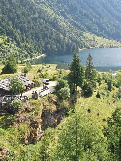 Eine malerische Landschaft mit einem See und einem Bauernhaus umgeben von Bäumen. Die Berge im Hintergrund vervollständigen das idyllische Bild. | © Herbert Raffalt